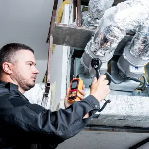 A technician uses a wrench and a digital meter to inspect HVAC ductwork covered in insulation material.