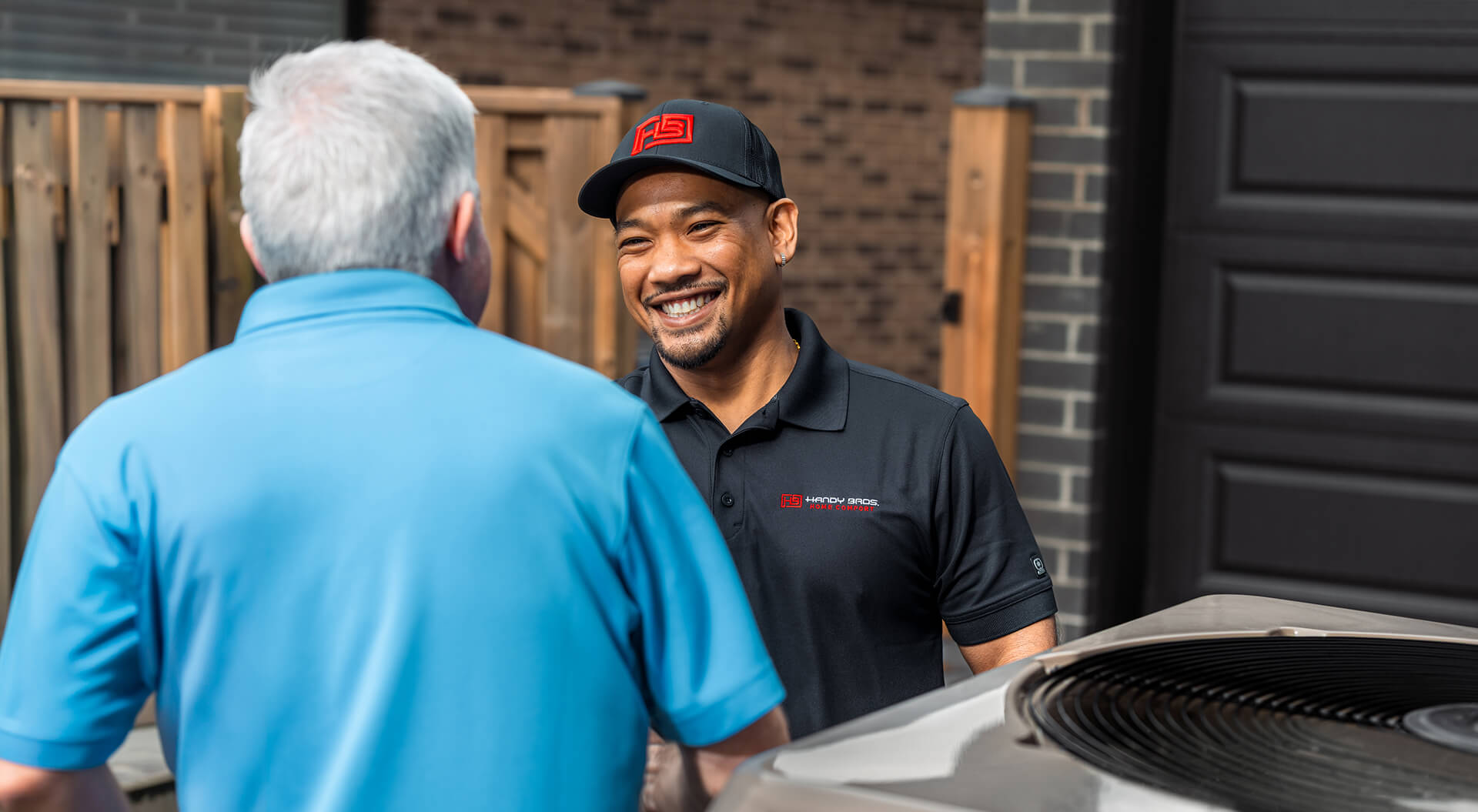 An HVAC technician in uniform smiles and talks with a customer outdoors in Windsor, next to an air conditioning unit during an AC repair.