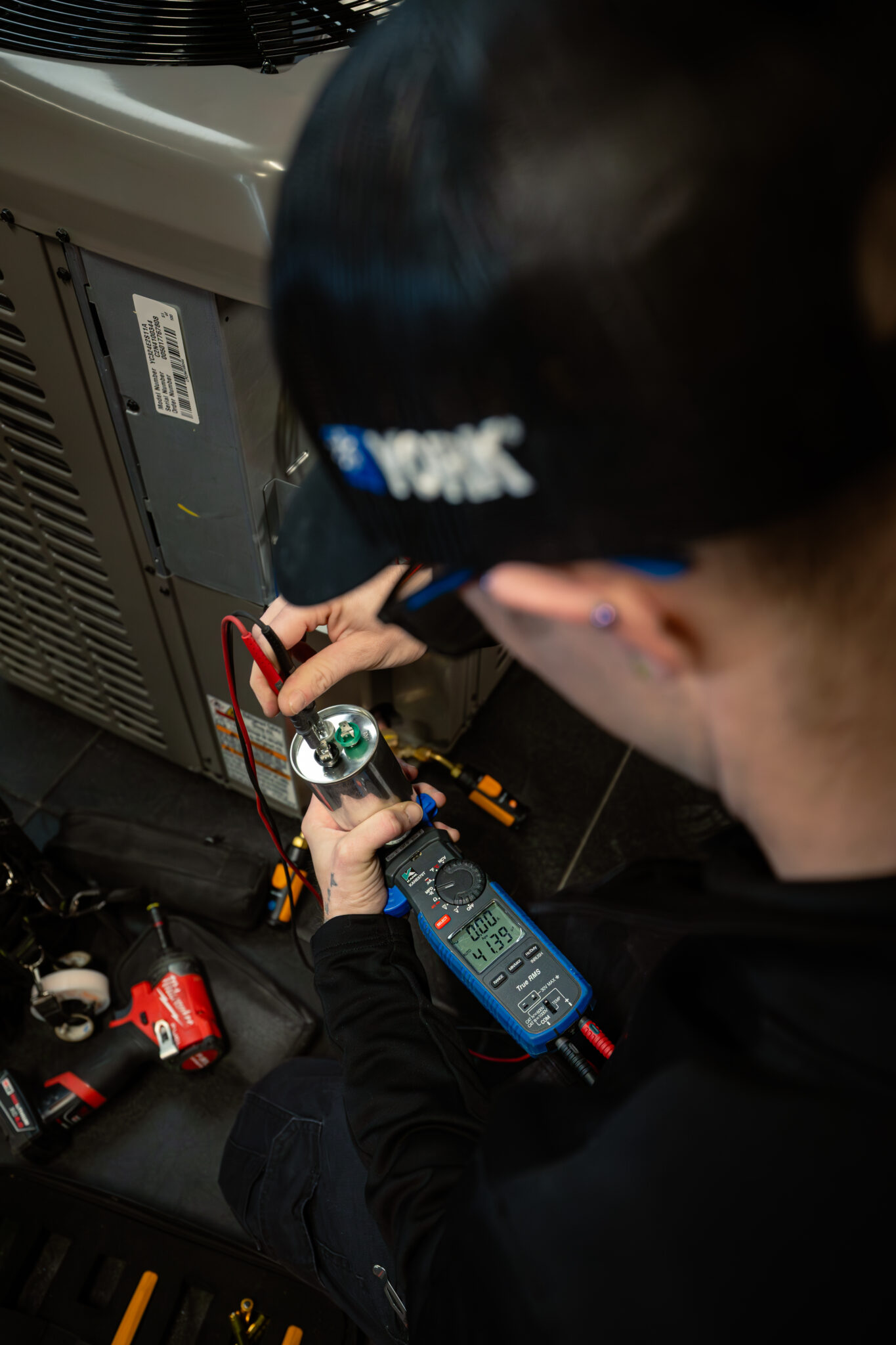 A technician uses a digital multimeter to measure a capacitor near an HVAC unit, checking for potential leaks during residential AC refrigerant installation and holding test probes on the capacitor terminals.