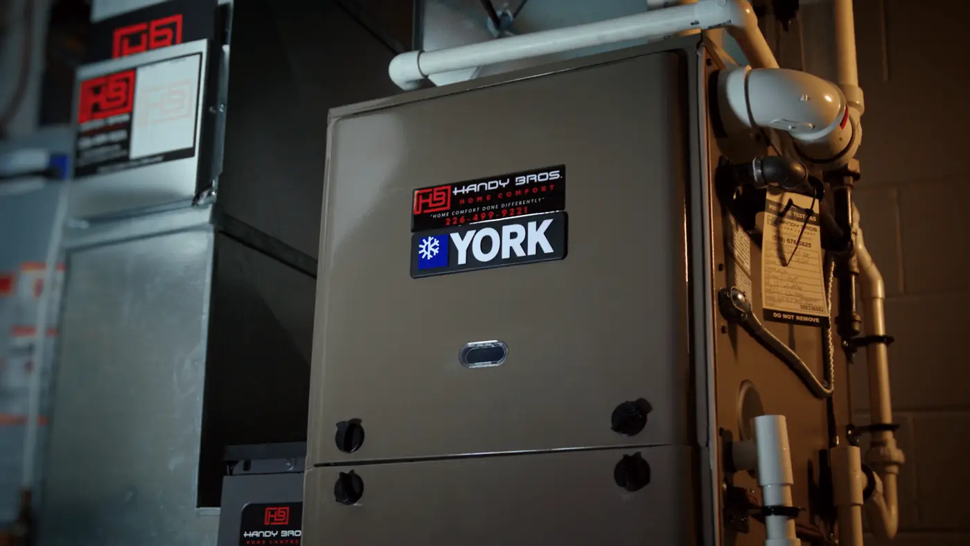 4k-graded-p4-furn-installed-pp00000000-mov-00-00-08-13-still001-hermes-nonmp4-1400 - handy bros. home comfort - handy bros. home comfort York-brand furnace in a utility room with exposed pipes and a Handy Bros sticker.