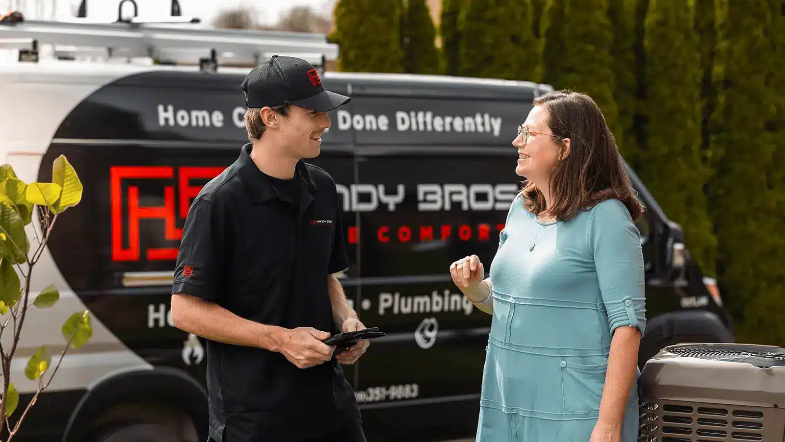 A technician in black polo and cap chats with a smiling woman beside a black service van with red logo and text in an outdoor driveway.
