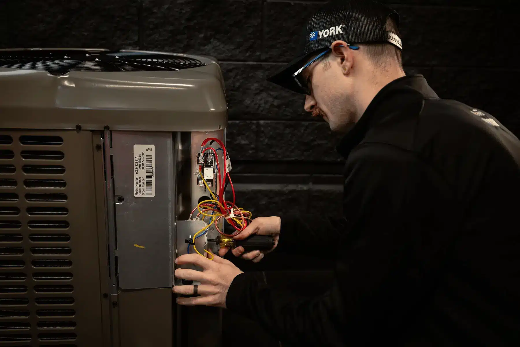 HVAC technician wiring the inside of an outdoor condenser unit, connecting multicolored wires to a control board.