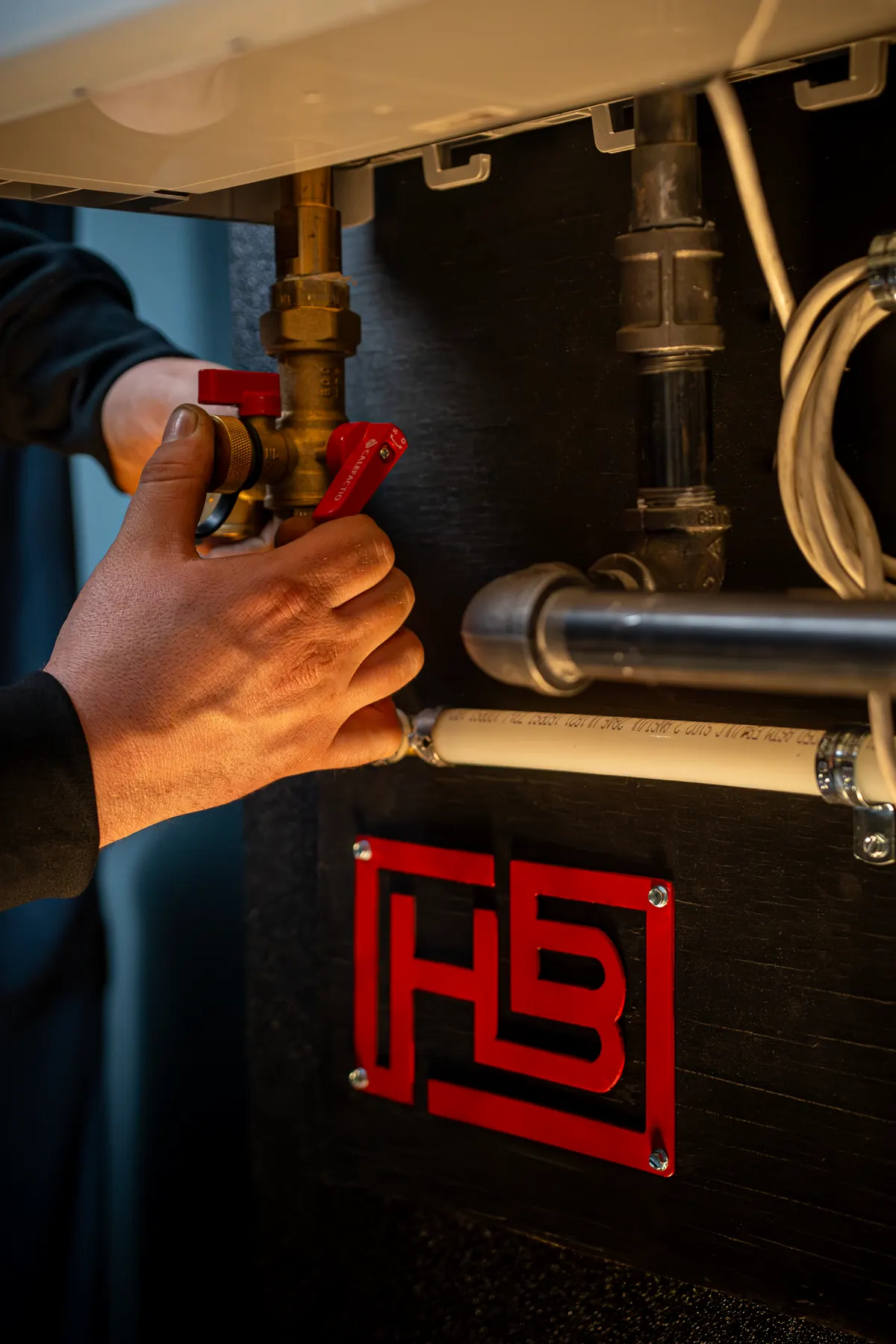 hand-tankless-gas-knob-1-optimized-1800 - handy bros. home comfort - handy bros. home comfort Close-up of a technician turning a red lever valve on a copper pipe under an industrial cabinet.
