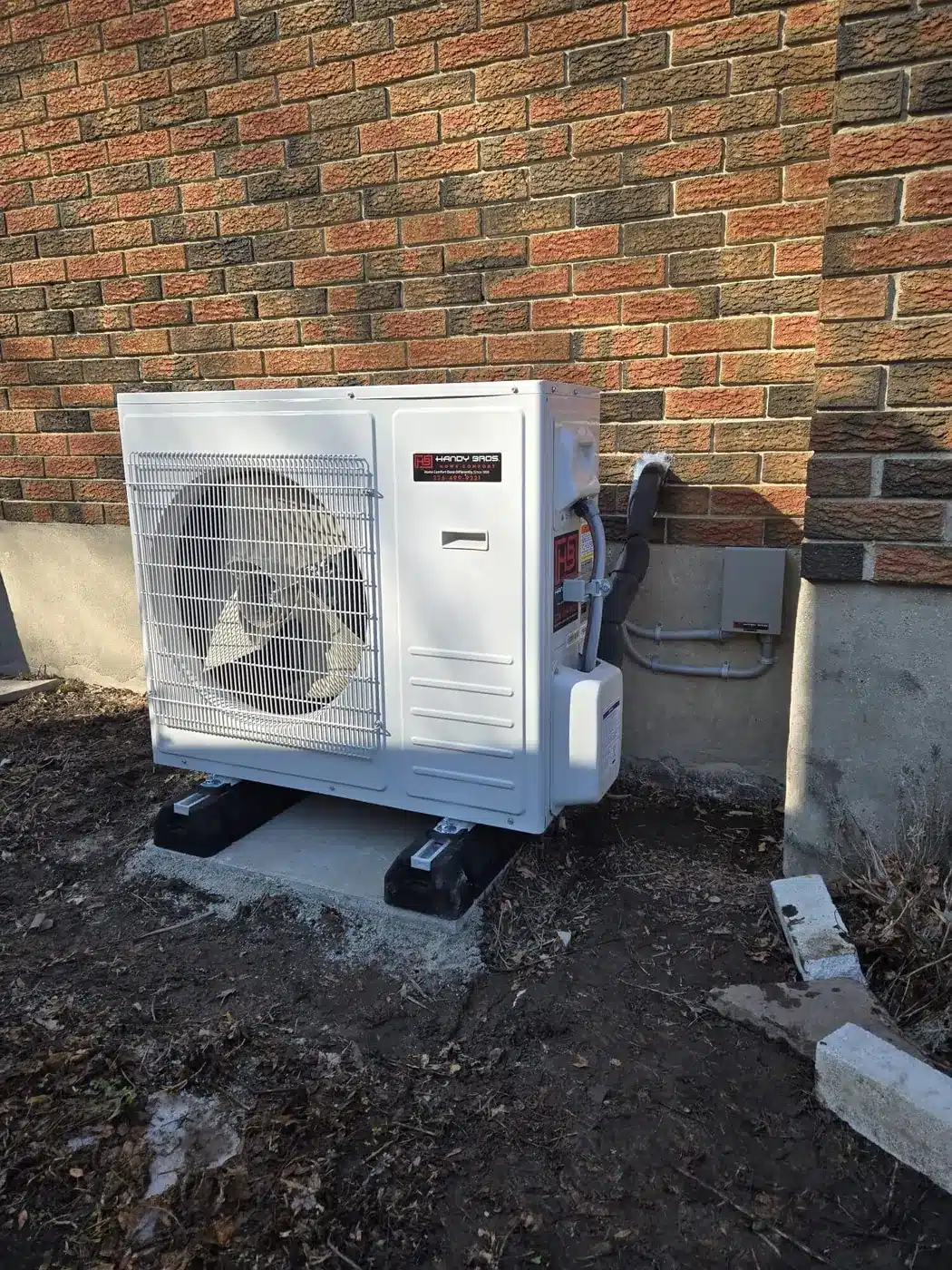 Outdoor air conditioner condenser unit on a concrete pad beside a brick wall, with a protective grille and attached refrigerant lines.