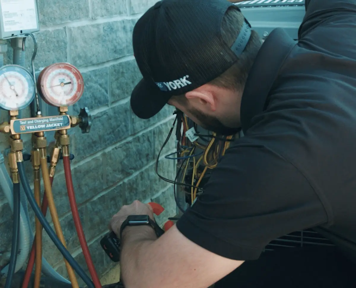 screenshot-2024-08-28-at-12-18-55-pm-e1738860055626-hermes-nonmp4-1400 - handy bros. home comfort - handy bros. home comfort HVAC technician crouching beside a refrigerant manifold with colorful hoses connected to a wall-mounted system outside.