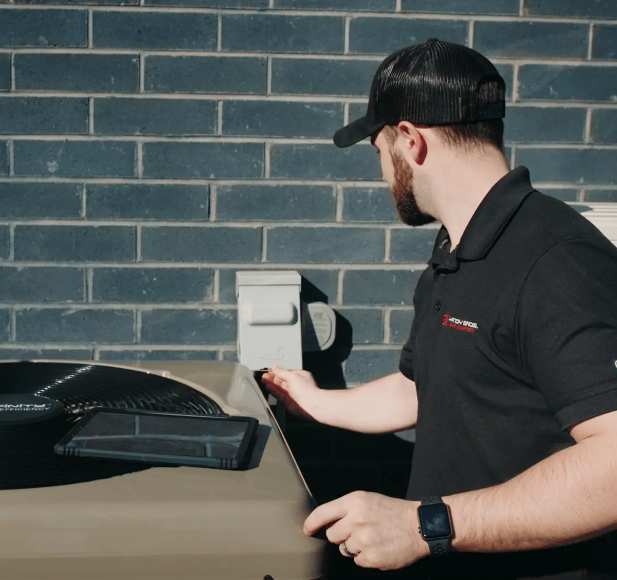 take-your-top-off-still-1-5-1-e1738858852968-hermes-nonmp4-1400 - handy bros. home comfort - handy bros. home comfort Technician in a black polo and cap services an outdoor HVAC condenser against a blue brick wall, with a tablet resting on the unit nearby.