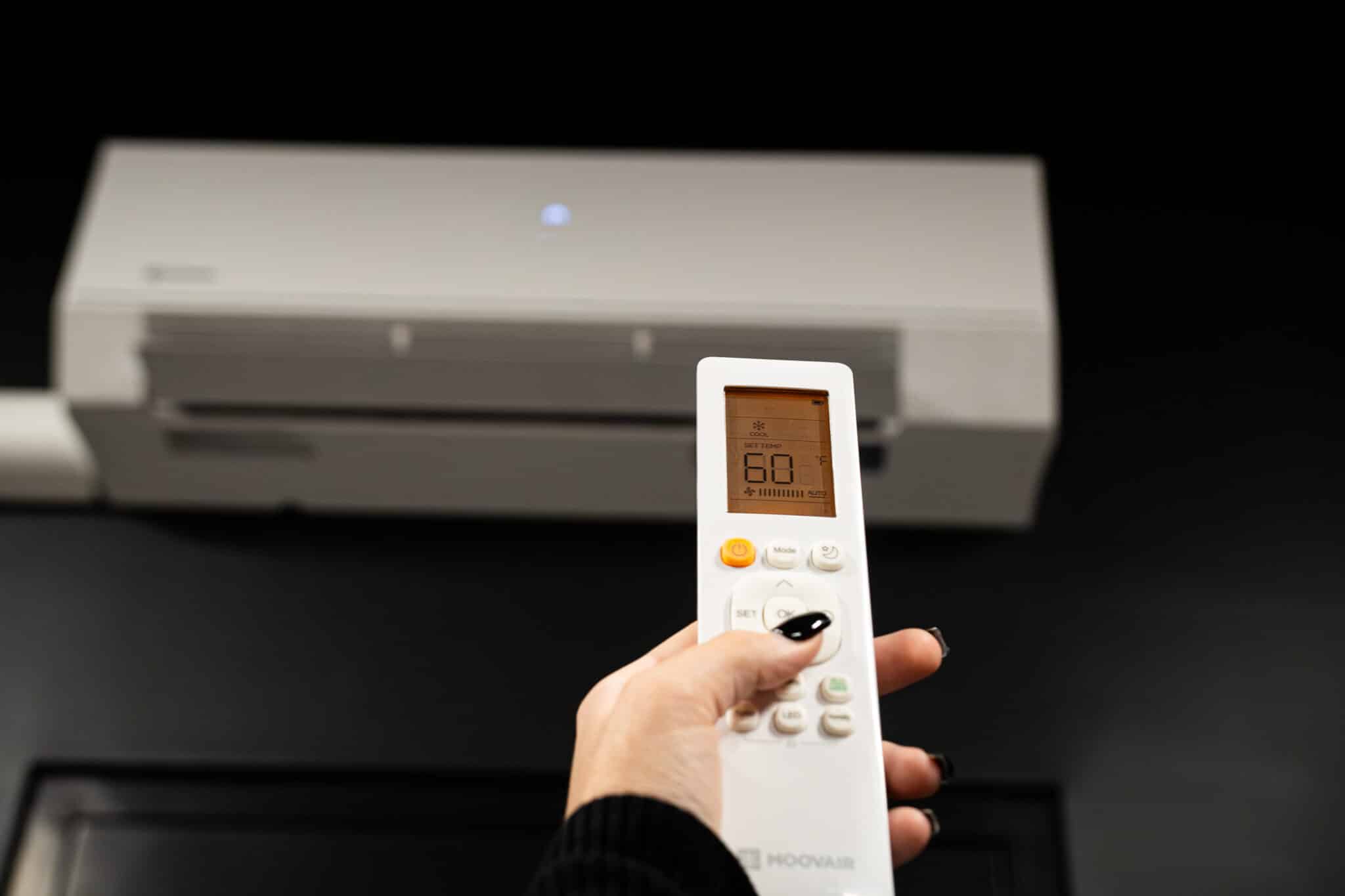 Person holding a white remote control pointed at a wall-mounted air conditioner, displaying 60°F on the screen.