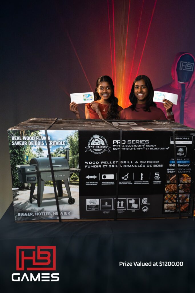 Two smiling women hold prize envelopes in front of a boxed Pit Boss grill at a games event.