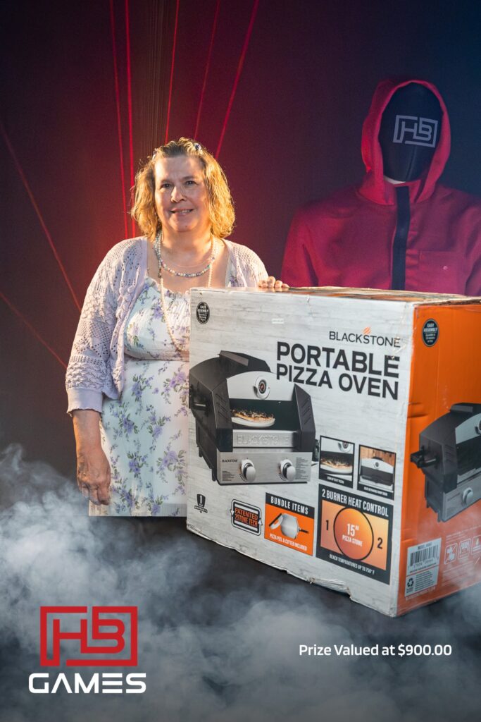 Smiling woman stands beside a boxed Blackstone portable pizza oven prize on a stage with a red-hooded figure and fog in the foreground.