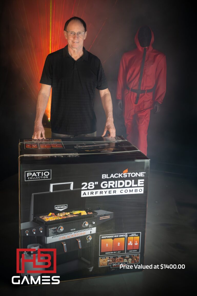 Man wearing a black polo stands beside a boxed Blackstone 28" griddle air fryer combo at a display event, with red laser lights in the background and a red-suited mannequin nearby.
