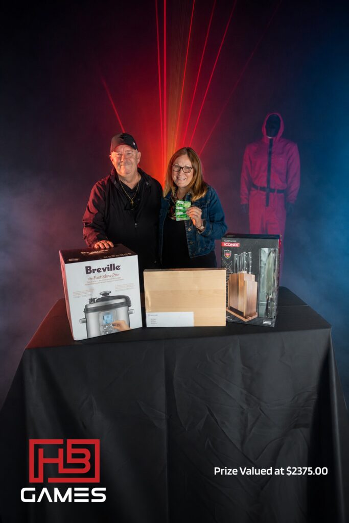 Two people smile behind a prize table with boxed appliances and prizes on display, red laser lights in the background.